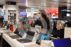 Young woman in medical face mask choosing laptop in tech store during Covid-19 pandemic