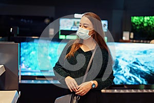 Young woman in medical face mask pensively examines variety of tv sets on display in tech store