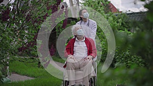Young woman in medical gown and coronavirus face mask rolling wheelchair with disabled old man in nursing home garden