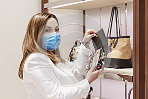 Young woman in a medical mask chooses a bag in the store. Shopping during the coronavirus pandemic