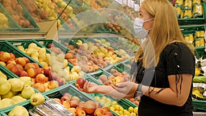 A young woman in a medical mask chooses fresh fruit in a grocery store.