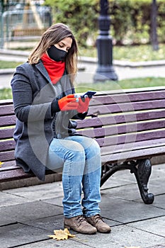 A young woman in a medical mask communicates online chatting on a mobile phone, sitting on a bench in an autumn park. Social