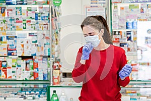 A young woman in a medical mask and gloves coughs, covering her face. In the background, the shop Windows of pharmacy products.