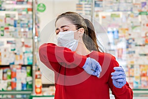 A young woman in a medical mask and gloves coughs, covering her face with the crook of her elbow. In the background, the shop