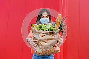 Young woman in medical mask holds an eco paper bag with food, fruits and vegetables, pepper, baguette, lettuce, safe online smart