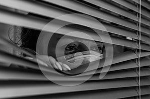 Young woman in medical mask looks out the window through the blinds