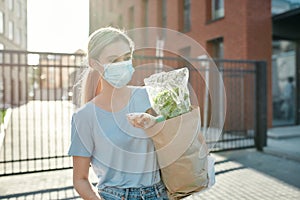 Young woman in medical mask and protective gloves walking with grocery bag to her car after doing shopping at a