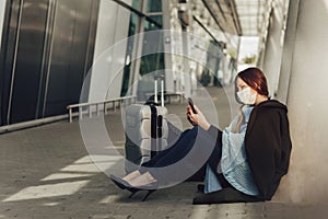 Young woman in medical mask sits near luggage in airport. Woman waiting for flight, using a cell phone and