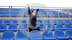 Young woman in medical mask sitting on stadium bleachers alone and rooting for her favorite sports team. Adult female