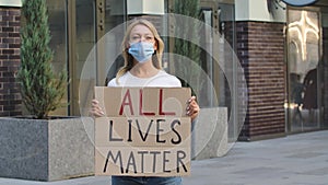 Young woman in medical mask stands with a cardboard poster ALL LIVES MATTER in a public place outdoor. No racism but