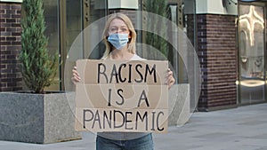 Young woman in medical mask stands with a cardboard poster RACISM IS A PANDEMIC in a public place outdoor. No racism