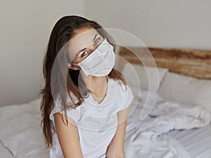 young woman in medical mask tilted her head to the side and sits on the bed indoors