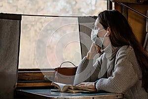 A young woman in medical mask is traveling by train alone and looking in window