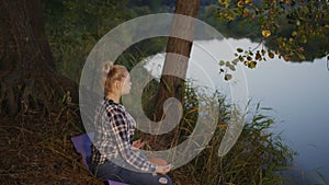 Young woman meditating in lotus pose on river shore in evening forest. Girl practicing yoga meditation on river coast