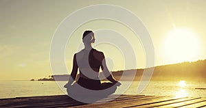 Young woman is meditating in Lotus Pose sitting on pier near the sea at sunset.