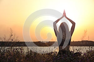Young woman meditating near river at sunset, back view