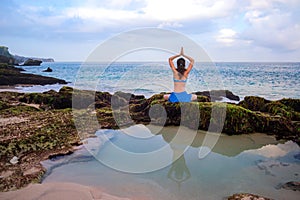 Young woman, meditating, practicing yoga and pranayama at the beach. Sunrise yoga practice. Hands raising in namaste mudra. View