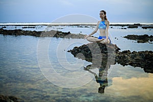 Young woman, meditating, practicing yoga and pranayama at the beach. Sunset yoga practice. Hands in gyan mudra. Water reflection.