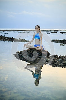 Young woman, meditating, practicing yoga and pranayama at the beach. Sunset yoga practice. Hands in gyan mudra. Water reflection.