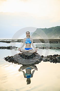 Young woman, meditating, practicing yoga and pranayama at the beach. Sunset yoga practice. Hands in gyan mudra. Water reflection.