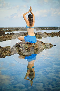 Young woman, meditating, practicing yoga and pranayama at the beach. Sunset yoga practice. Hands raising in namaste mudra. View