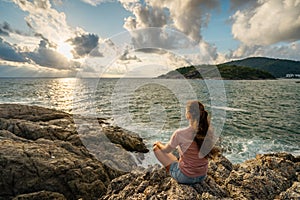 Young woman meditating at the seaside sitting in the lotus position on rocks overlooking the sea with sunset in a