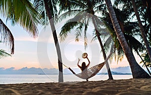 Young woman meeting sunrise sitting in hammock on the sand beach under the palm trees