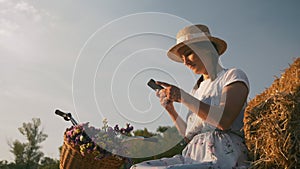 Young woman with mobile phone in hands is sitting on straw at sunset. Girl in hat and blue dress is typing on smartphone and relax