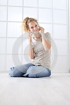 Young woman with mobile phone sitting on the floor dressed casual with curly and long red hair  on white window background