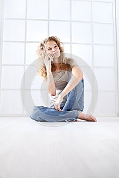 Young woman with mobile phone sitting on the floor dressed casual with curly and long red hair  on white window background