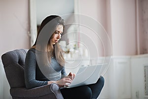 Young woman in modern luxury apartment, sitting comfortable in armchair holding computer on her laps, relaxing, working