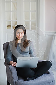 Young woman in modern luxury apartment, sitting comfortable in armchair holding computer on her laps, relaxing, working