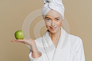 Young woman moisturizes face with fresh apple dressed in shite coat wrapped towel on head after taking shower