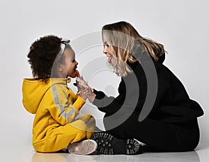 Young woman mother and curly dark-skinned daughter girl in sport jumpsuit and sneakers sit cross-legged face to face