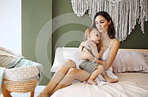 Young woman mother expressing love to little baby, sitting on bed with sleeping child in hands and kissing it in forehead