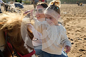 Young woman mother and her adorable little kid girl, a lovely daughter smiling and stroking the pony