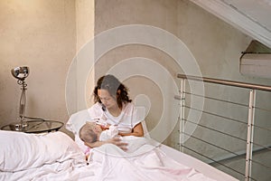 Young woman mother holding and breastfeeding her newborn baby, sitting on the bed in white bedchamber interior