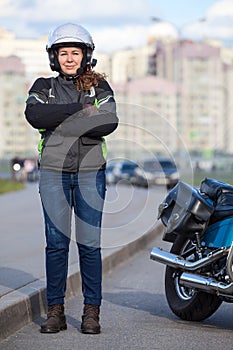 Young woman motorcyclist standing roadside near her motorcycle, full length portrait with cross arms on breast