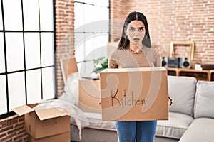 Young woman moving to a new home holding box in shock face, looking skeptical and sarcastic, surprised with open mouth