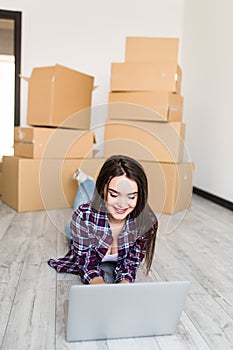 Young woman is moving using a laptop and smiling, lying among cardboard boxes in new home