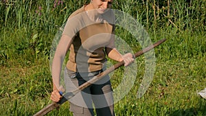 Young woman mowing the green grass with the scythe on the summer field by the river. Beautiful landscape. Work in the
