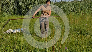 Young woman mowing the green grass with the scythe on the summer field by the river. Beautiful landscape. Work in the