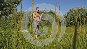 Young woman mowing the green grass with the scythe on the summer field by the river. Beautiful landscape. Work in the