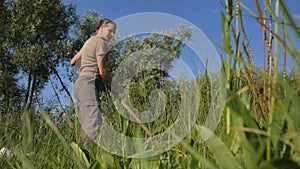 Young woman mowing the green grass with the scythe on the summer field by the river. Beautiful landscape. Work in the