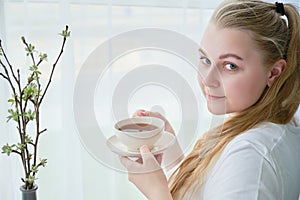 Young woman with a mug of tea in her hands sits near the window, space for text
