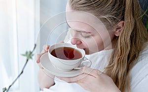 Young woman with a mug of tea in her hands sits near the window, space for text