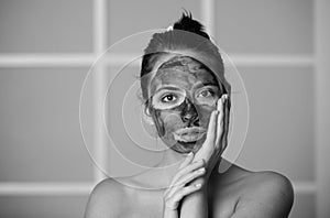 Young woman with naked shoulder and clay mask on face. Beautiful model applying cosmetic cream treatment on face. Facial