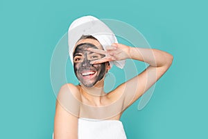 Young woman with naked shoulder and clay mask on face, on blue. Beautiful model applying cosmetic cream treatment on