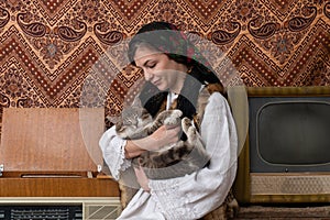 Young woman in national closes sitting among aged retro room and holding cat in her hands