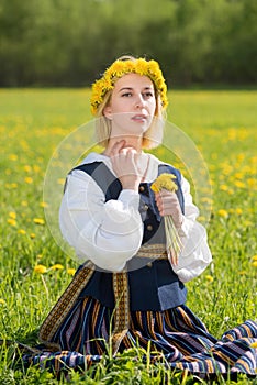 Young woman in national clothes wearing yellow dandelion wreath in spring field. Ligo